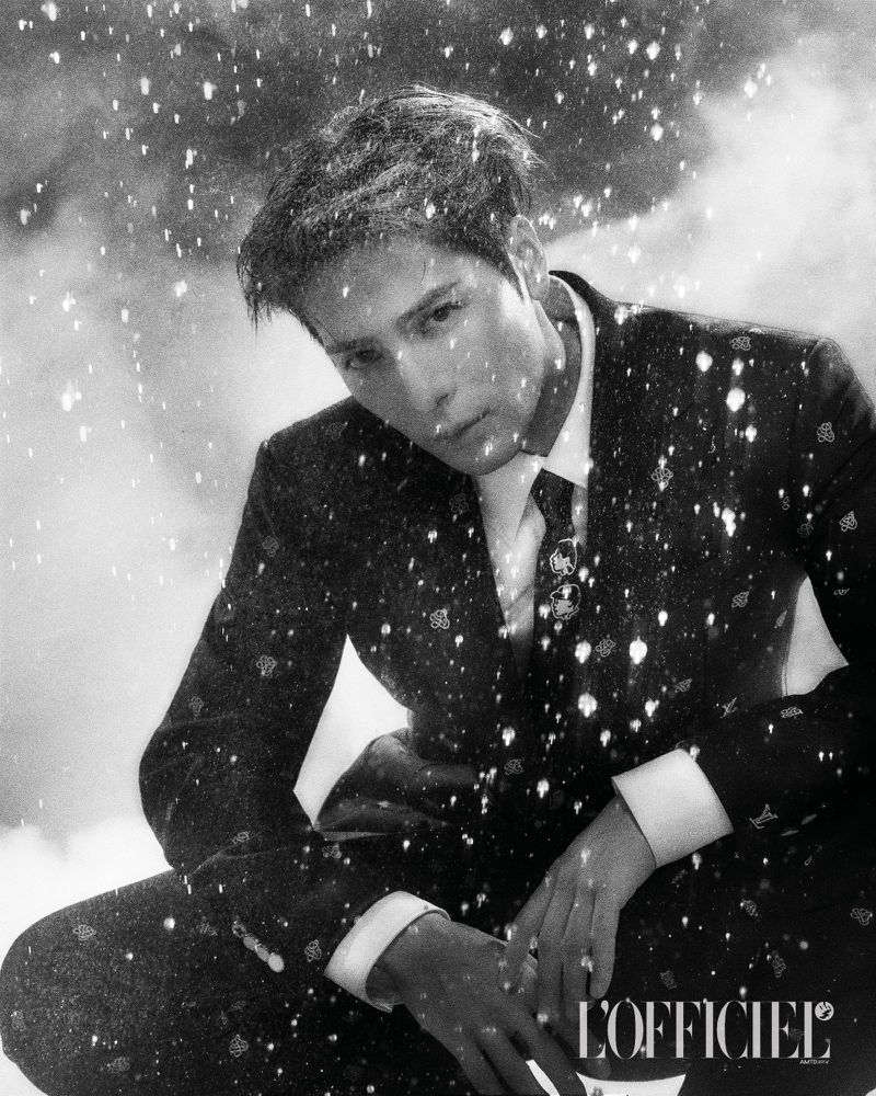 Monochrome studio portrait of an adult male model in a patterned suit, gazing downward against a snowy background with the L'Officiel logo.