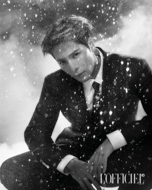Monochrome studio portrait of an adult male model in a patterned suit, gazing downward against a snowy background with the L'Officiel logo.