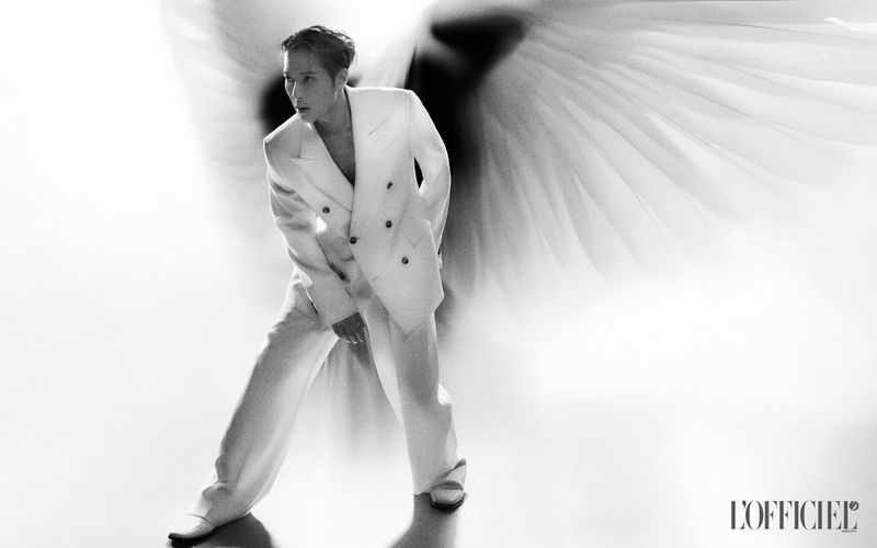 Monochrome portrait of an adult male fashion model in a white suit jacket and pants, standing in front of large white feathers.