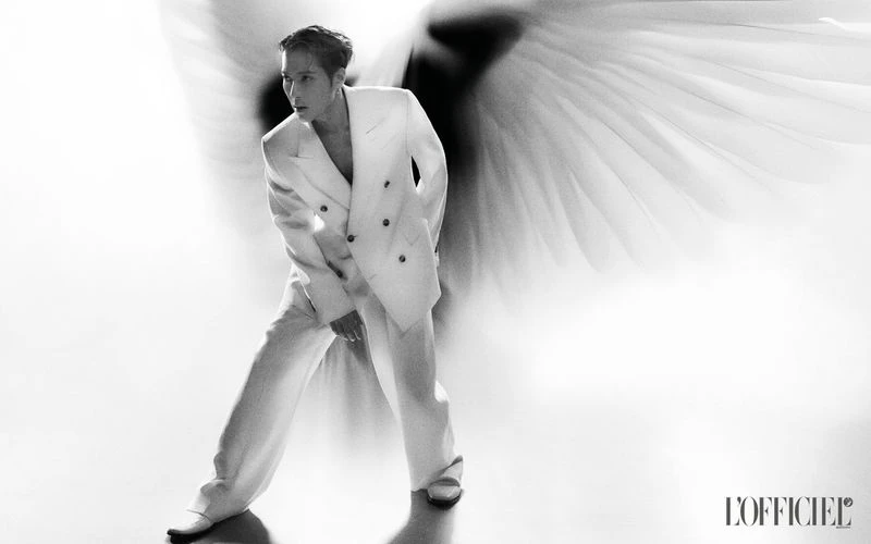 Monochrome portrait of an adult male fashion model in a white suit jacket and pants, standing in front of large white feathers.