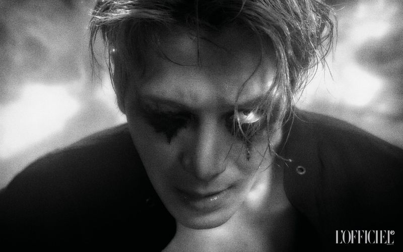 Black and white portrait of a male model with dramatic black eye makeup looking downward against a blurred studio background.
