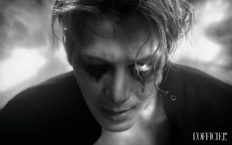 Black and white portrait of a male model with dramatic black eye makeup looking downward against a blurred studio background.