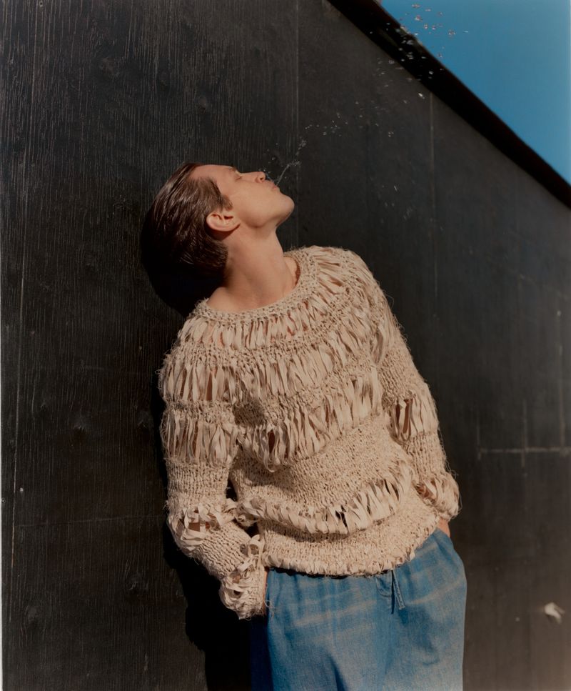 An adult is shown from a low angle leaning back against a dark wall and spitting water upwards into the air in an outdoor setting.
