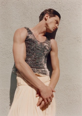 Studio shot of a muscular adult male model wearing a metal tank top and flowing skirt against a textured wall in soft, diffused lighting.