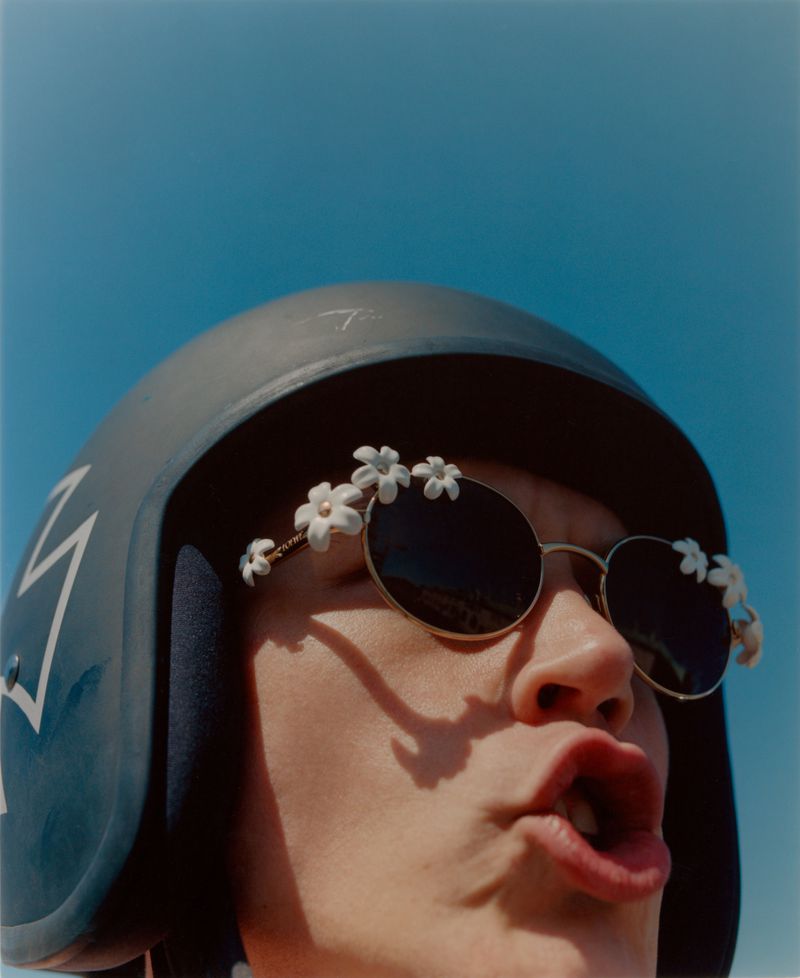 Close-up portrait of a person wearing a helmet, round sunglasses with flower accents, with a blue sky in the background.