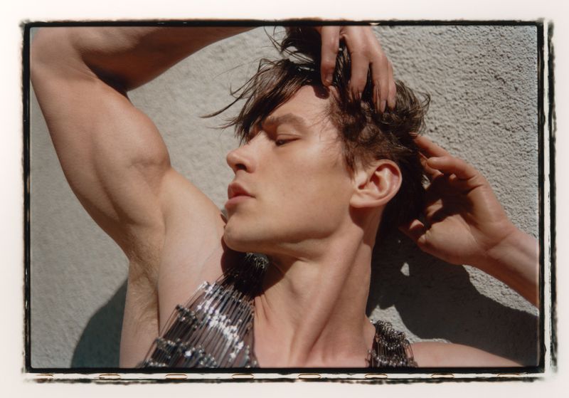 Close-up studio shot of a young adult male model with hands in his hair, wearing a metallic mesh top against a textured gray wall.