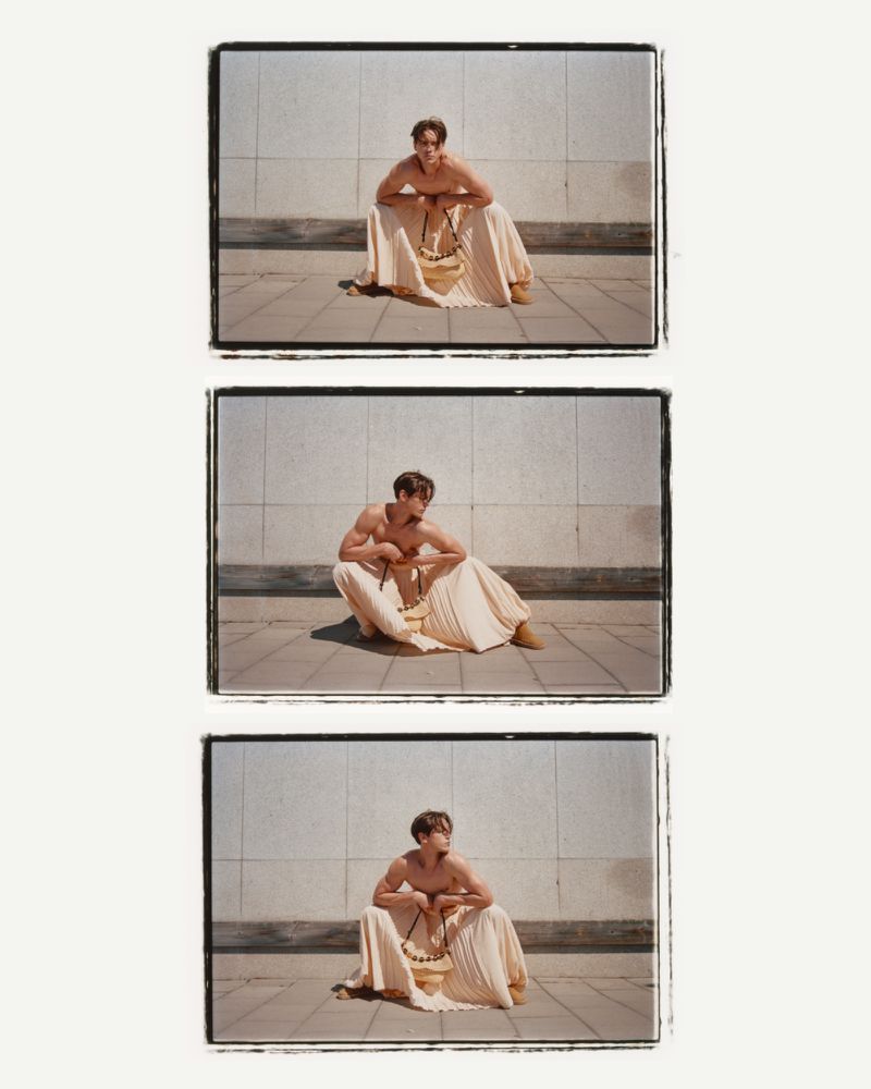 Three film-strip style frames show a shirtless male model sitting outside, wearing a tan skirt and beaded necklace, posing against a wall.