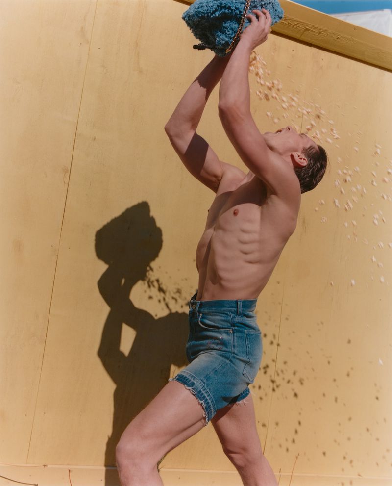 A shirtless man stands against a yellow wall, holding a blue textured bag over his head, with pale contents falling around him.