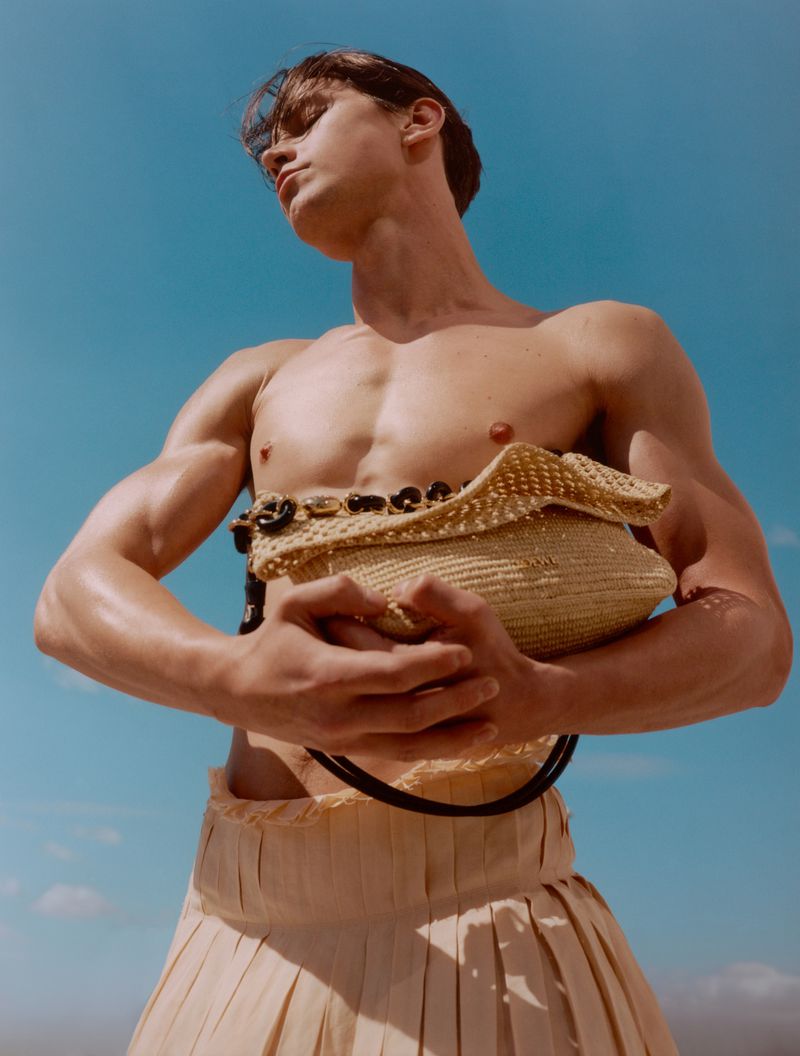 Shirtless man with a woven bag posing against a blue sky backdrop, showcasing a unique blend of fashion and natural elements.