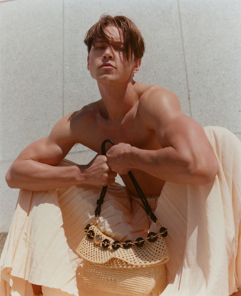 Fashion editorial shot of a shirtless man holding a straw handbag while draped with a beige garment outdoors in bright sunlight.