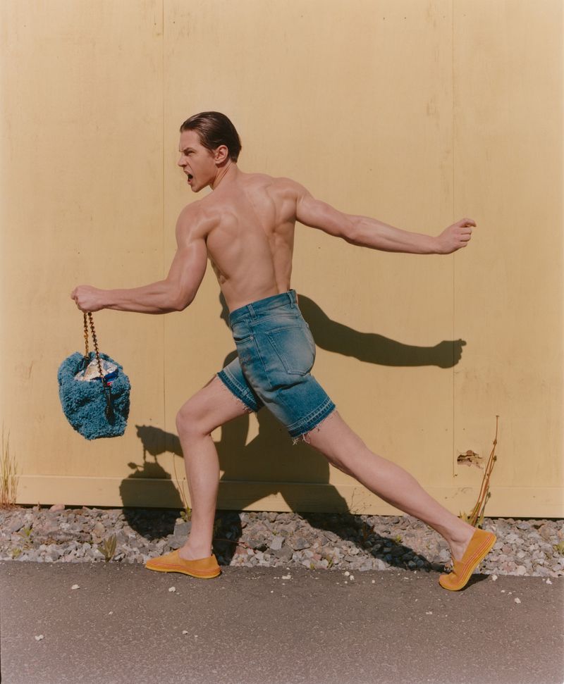 An athletic, shirtless adult male runs outdoors holding a blue crocheted bucket bag, wearing jean shorts and yellow shoes against a yellow wall.