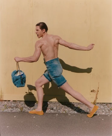 An athletic, shirtless adult male runs outdoors holding a blue crocheted bucket bag, wearing jean shorts and yellow shoes against a yellow wall.