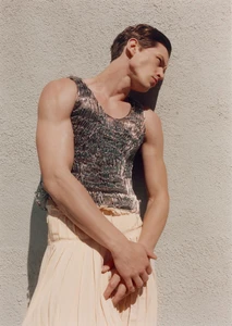 Man wearing a textured metallic tank top leaning against a light textured wall in sunlight