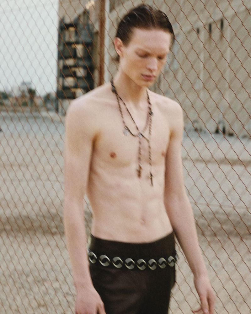 Shirtless male model poses in a gritty urban environment with a chain-link fence and aged buildings visible in the background on a sunny day.