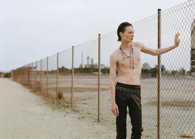 A slender, shirtless man stands beside a chain-link fence in a stark, outdoor environment on a cloudy day, showcasing dark pants and beaded necklaces.