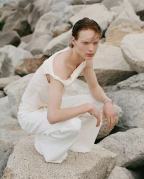 Young man with wet reddish hair squatting on rocky coast wearing white top and trousers for IVAN project