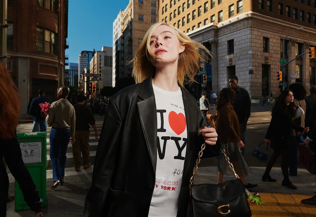 Elle Fanning poses on a city street wearing a black leather blazer, graphic t-shirt, and purse in an outdoor fashion shot with pedestrians.