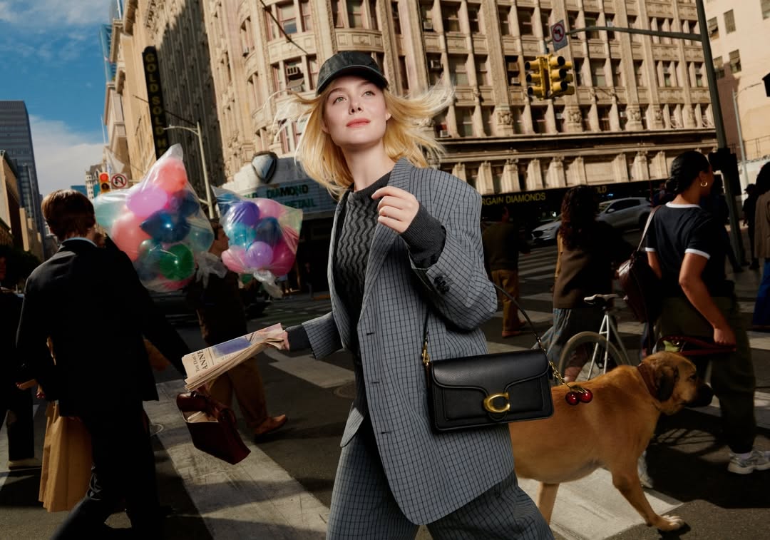 Elle Fanning walks across a city street in a grey suit, carrying a newspaper with a Coach bag, accompanied by a dog and balloons.