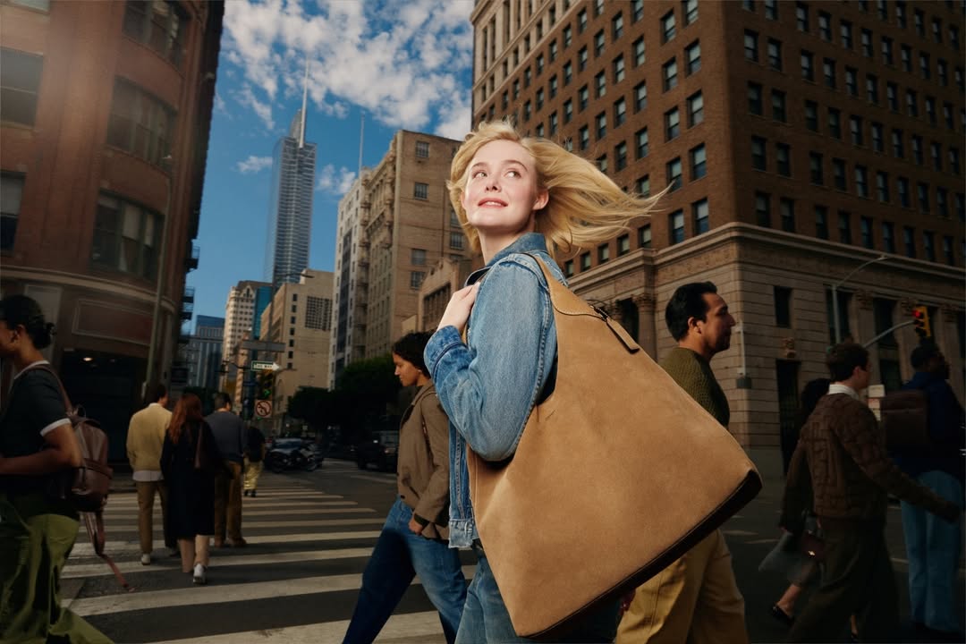 Woman crossing a street in a city with tall buildings. She has blonde hair and carries a large tan bag. Pedestrians surround her.