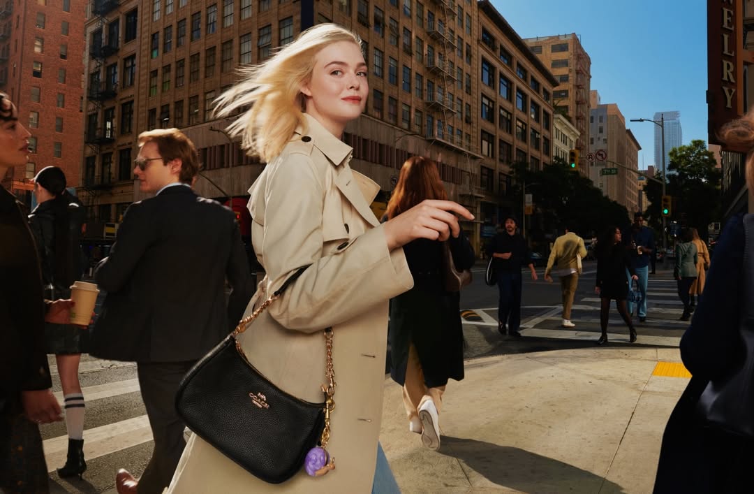Fashion shot of a woman with blonde hair, wearing a trench coat and carrying a black purse, walking on a city street crosswalk.