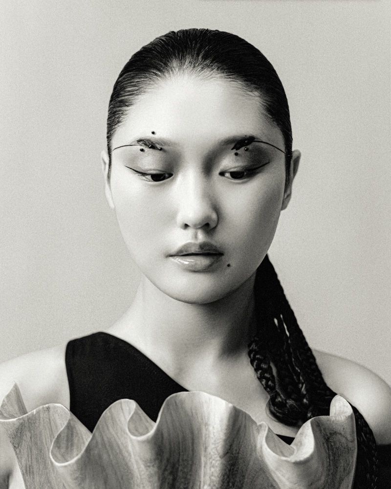 Close-up portrait of an Asian woman with artistic eye makeup and braided hair, wearing a black top and a ruffled collar.