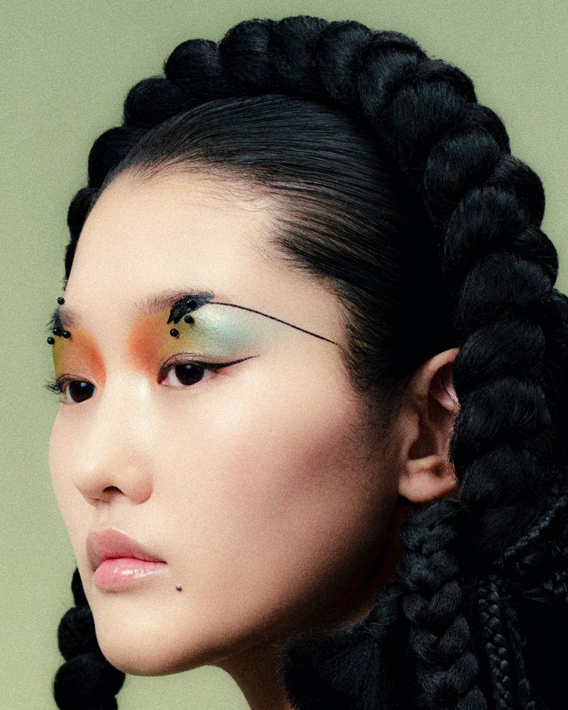Close-up portrait of a woman with creative makeup, featuring orange and green eyeshadow, black eyeliner, and a black braided hairstyle.