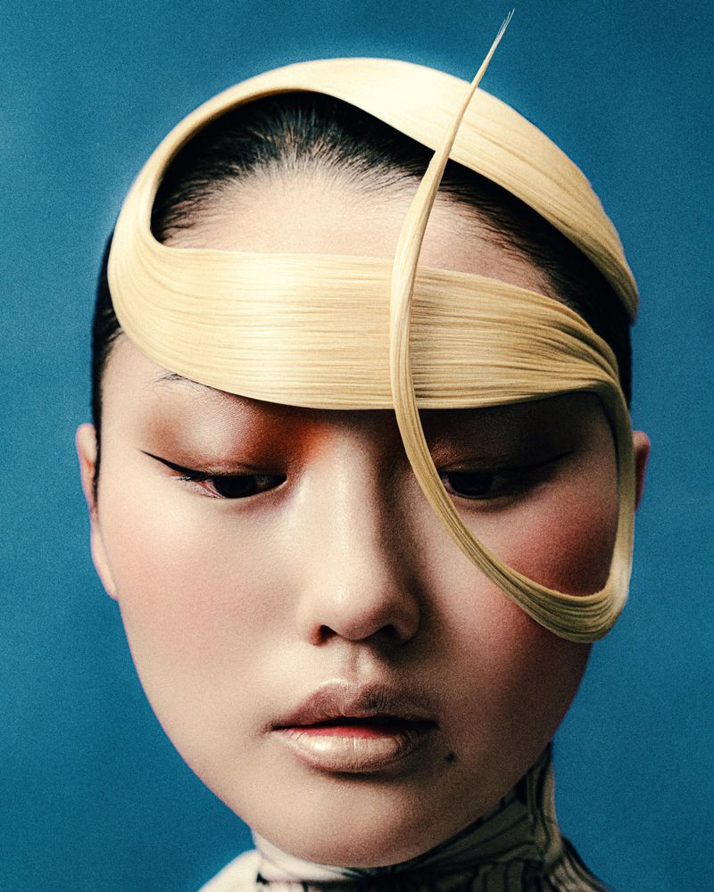 Headshot of an Asian woman with blonde hair styled as a head wrap in a studio portrait with heavy blush and graphic black eyeliner.