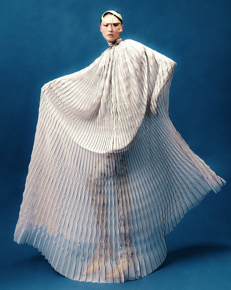 Fashion shot of a model in flowing pleated white gown against a blue backdrop, captured in a studio with professional lighting.