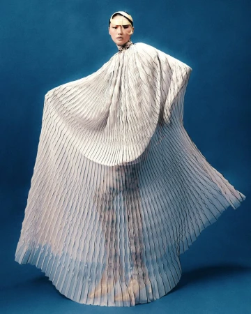 Fashion shot of a model in flowing pleated white gown against a blue backdrop, captured in a studio with professional lighting.