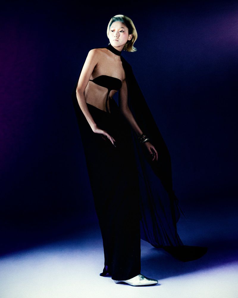 A woman poses in black formal wear with an open-back top and floor-length skirt against a dark blue and purple backdrop.