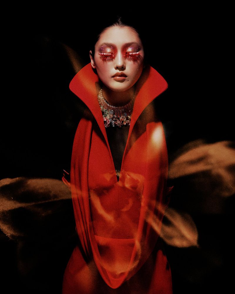 Portrait of a woman with dramatic red eye makeup and a large collar necklace, wearing a red gown, against a dark background in a studio.