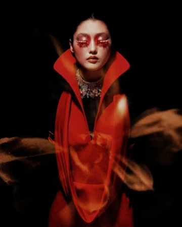 Portrait of a woman with dramatic red eye makeup and a large collar necklace, wearing a red gown, against a dark background in a studio.