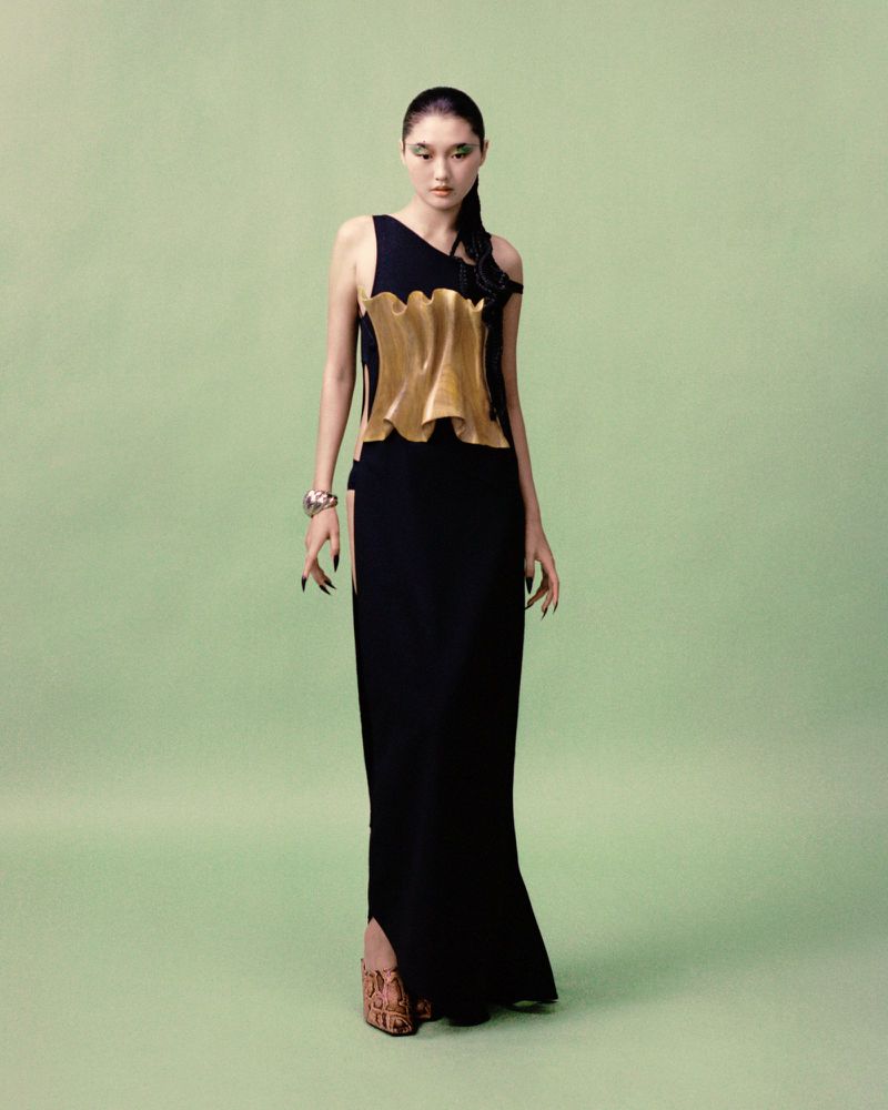 A slender Asian woman wearing a black gown with a gold-colored textured top is standing in a photo studio with a light-green backdrop.