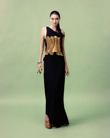 A slender Asian woman wearing a black gown with a gold-colored textured top is standing in a photo studio with a light-green backdrop.