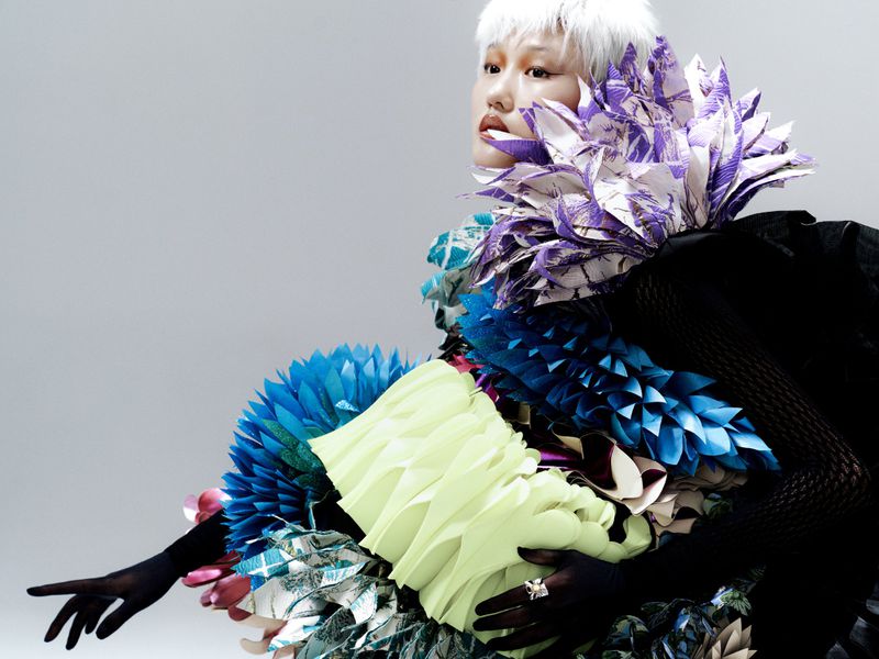 Elegant Asian woman wearing sculptural origami dress with black gloves poses against a grey backdrop in a striking studio shot.