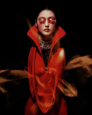 Model with dramatic red eye makeup and tall collar wearing an ornate necklace against a dark background