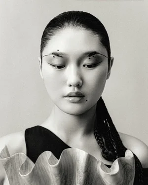 Portrait of an East Asian model with sleek dark hair and avant-garde makeup holding a sculpted object against a grey background.