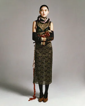 Model standing against a neutral studio backdrop wearing a patterned dress, long gloves, and holding a prop cluster.