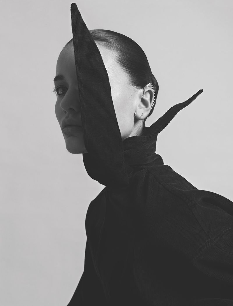 Monochrome portrait of a woman in high-fashion attire with a dramatic collar, gazing intently, set against a light backdrop.