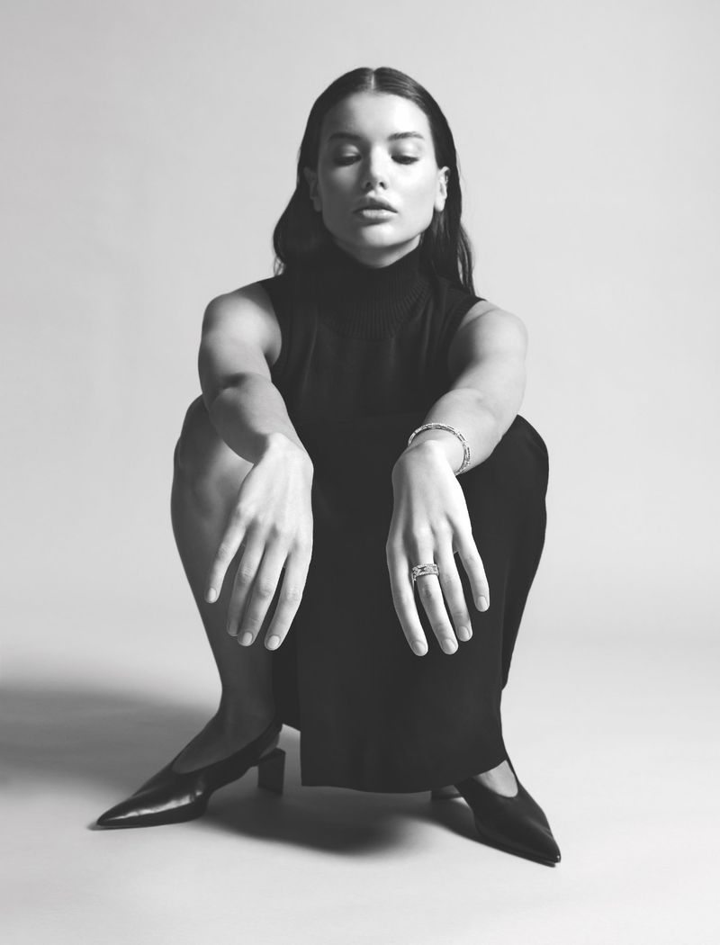 Woman in black dress and heels sitting with closed eyes in a studio, wearing bracelet and ring, black and white photography. 