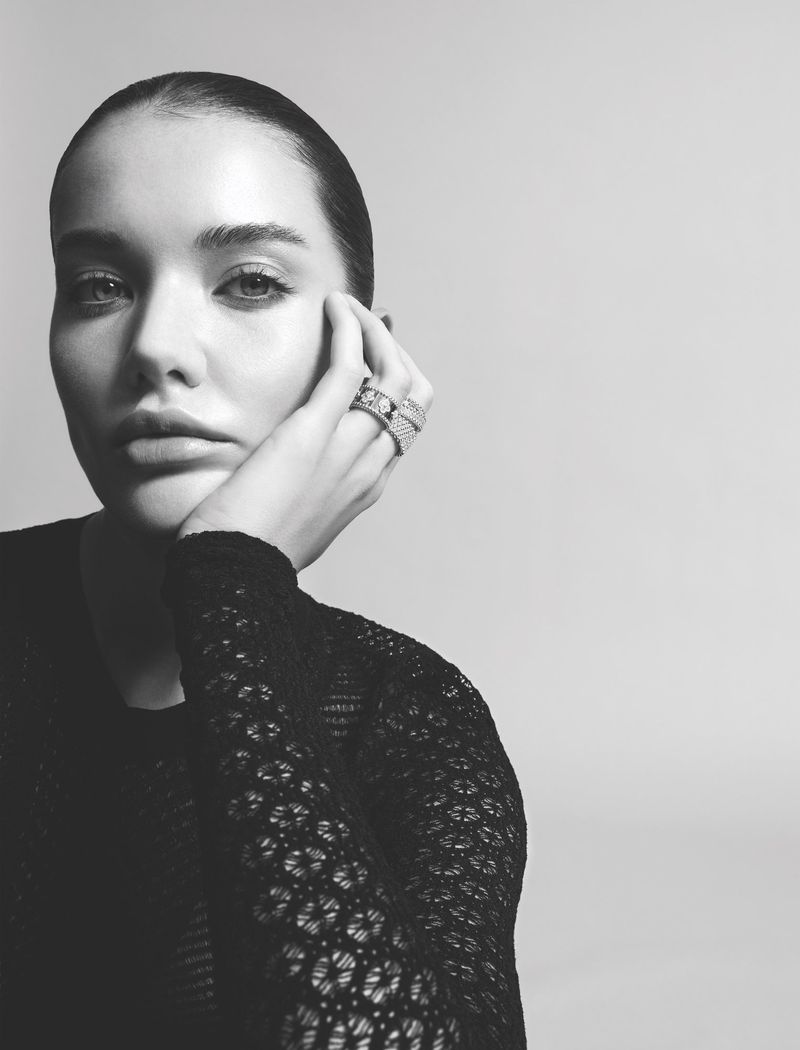 Portrait of a woman with her hand near her face, wearing statement rings and a black, patterned top against a plain, neutral backdrop.
