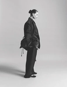 Model in black suit holding a dark leather handbag against a plain grey studio background