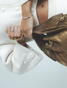 Close-up of a person wearing a white knit jacket holding a quilted metallic gold handbag against a white background.