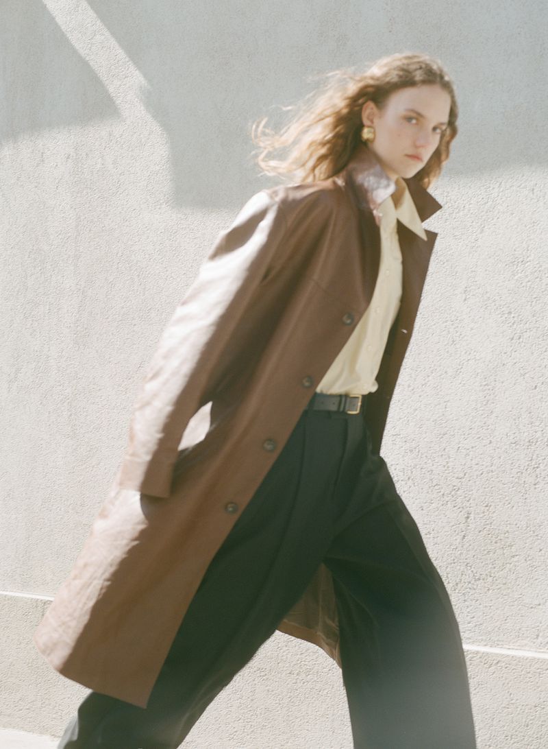 A stylish woman walks in a brown trench coat, black pants, and gold earrings, with a neutral concrete background and soft, warm lighting.