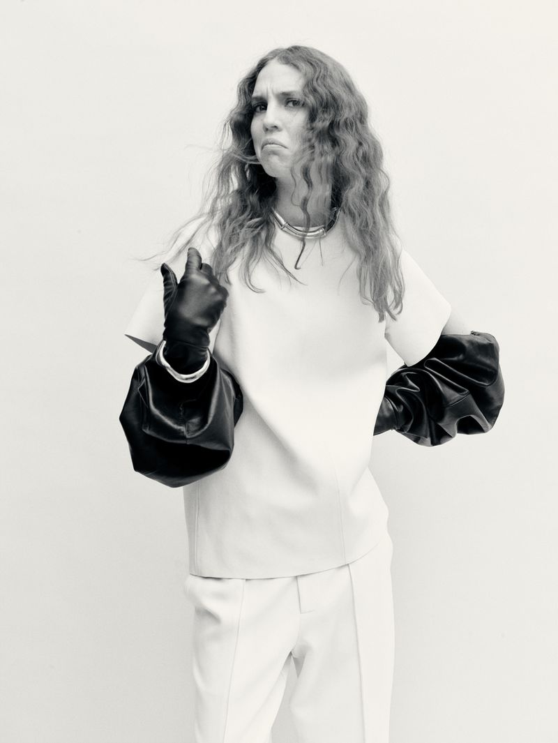 Portrait of a woman in a white suit and black gloves striking a pose in front of a white background, looking displeased.