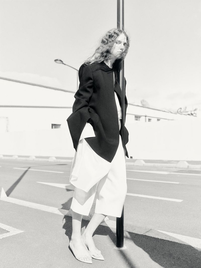 Black-and-white shot of a model in a black jacket and white culottes leaning against a pole in an urban outdoor environment on asphalt.