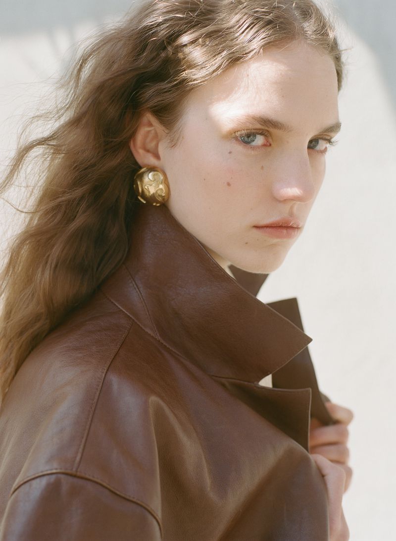Portrait of a young woman with golden earrings wearing a brown leather jacket against a pale background in natural diffused light.