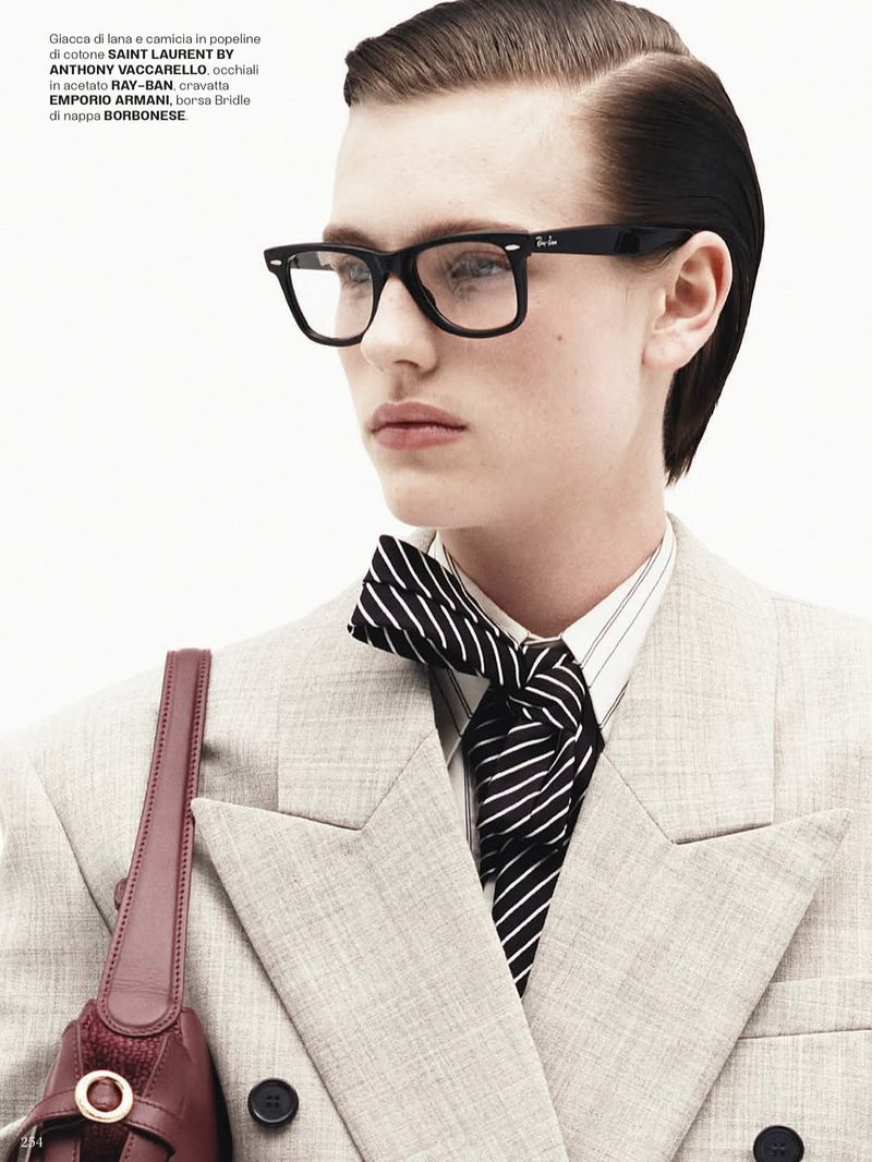 Fashion editorial shot features a young adult wearing a double-breasted suit, Ray-Ban glasses, and a striped bow tie against a white backdrop.
