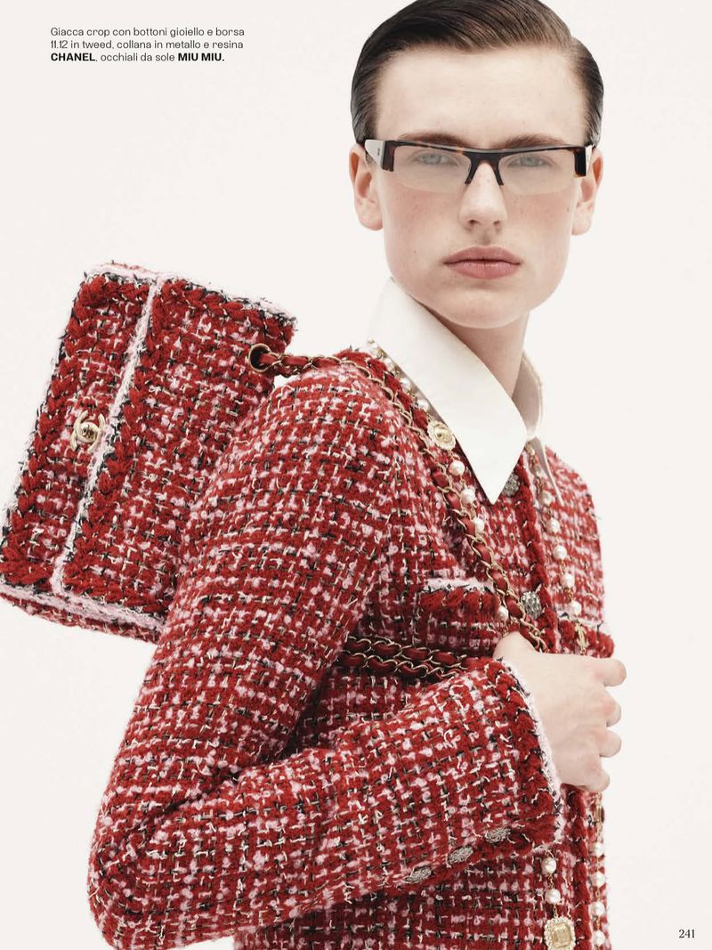 Fashion editorial shot of a stylish woman wearing a tweed Chanel suit, carrying a matching handbag, and wearing eyeglasses against a white backdrop.
