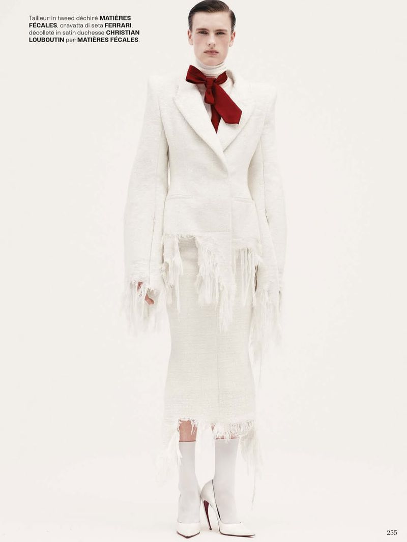 Full-length shot of a male model in a white tweed suit with fringe details and white heels, posing against a solid white backdrop.